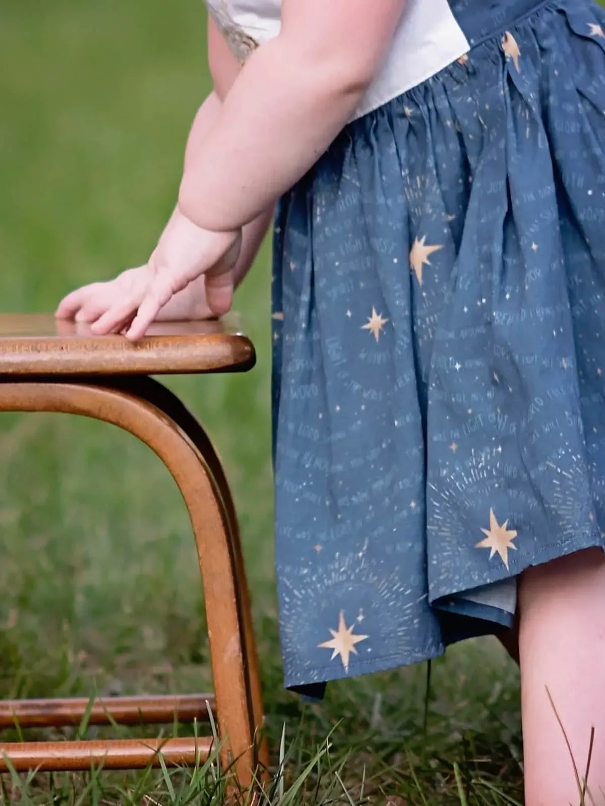 A child in a blue skirt with gold stars and text stands next to a wooden chair.