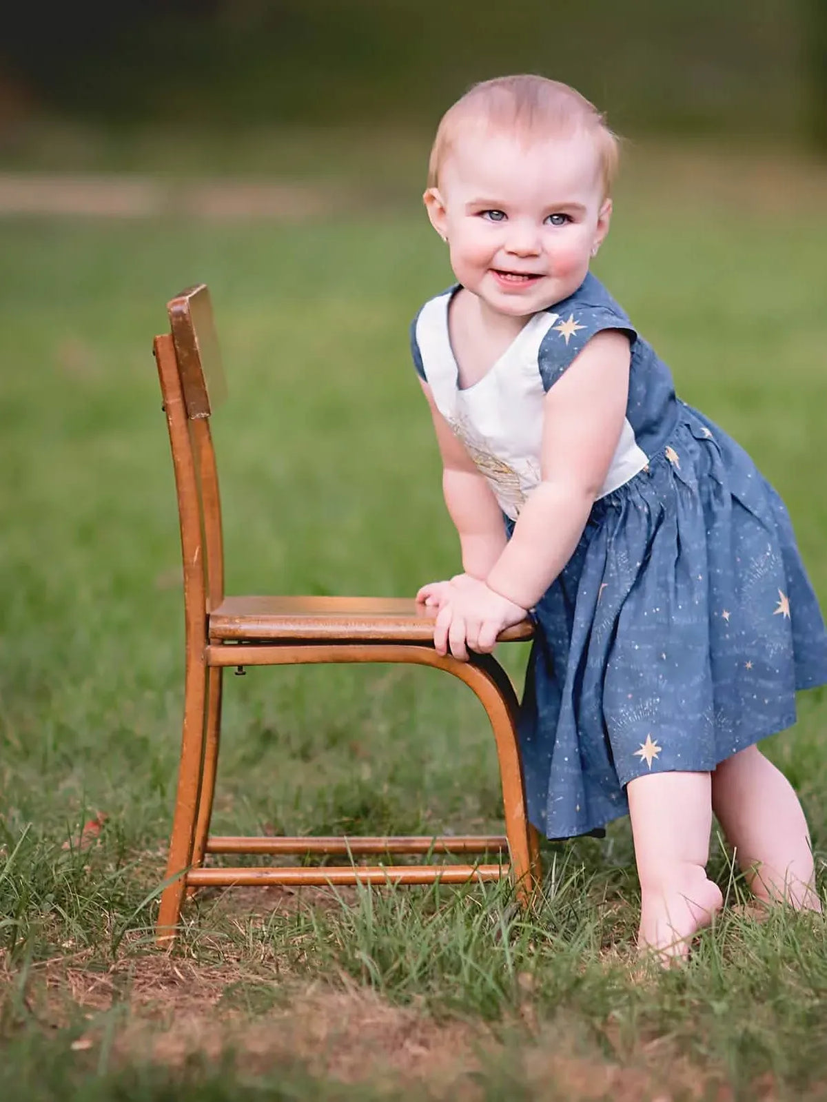 A smiling baby in a blue and white dress with gold stars leans on a wooden chair.