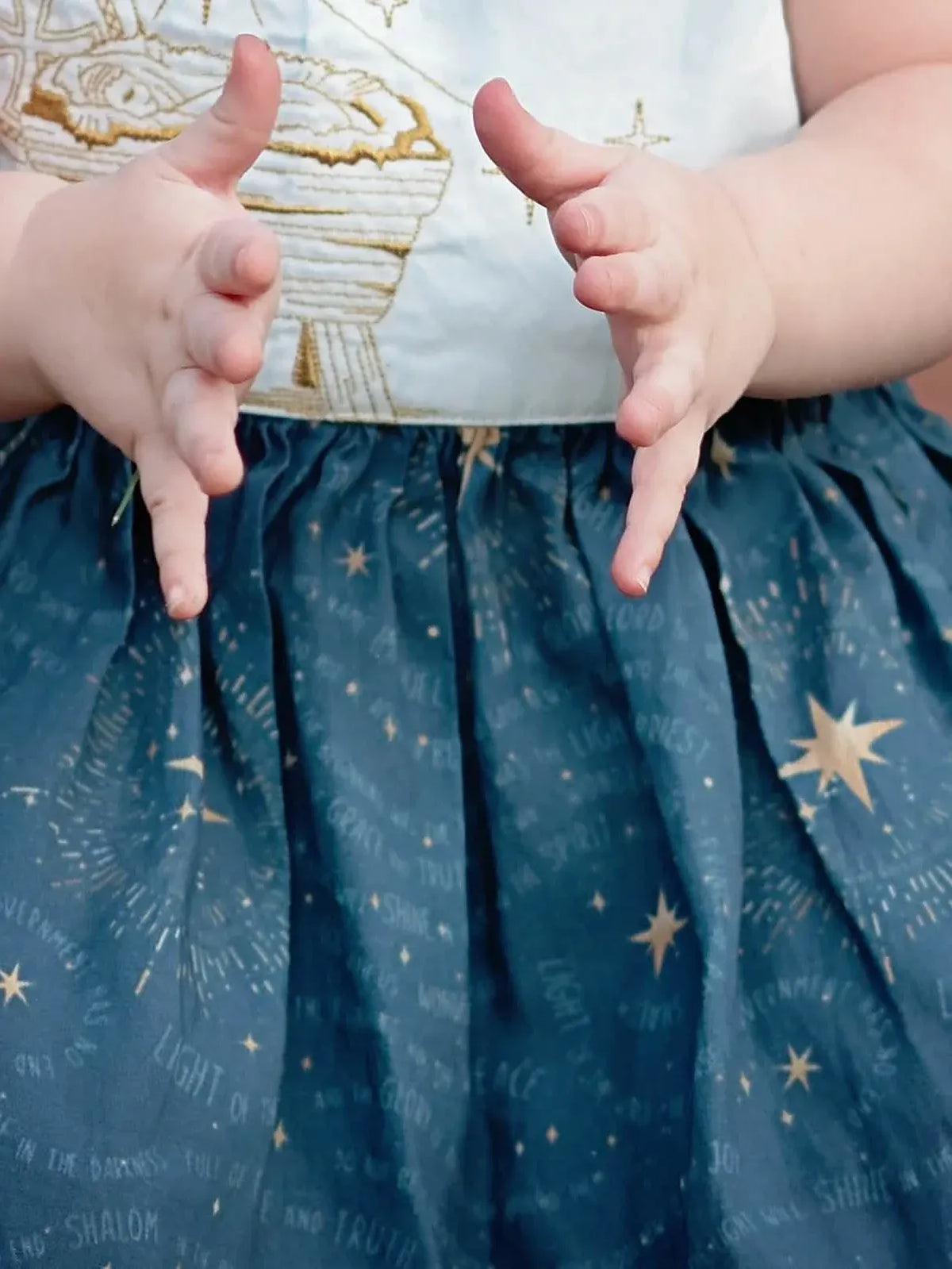 A baby's hands are held up over a blue skirt with gold stars and text.