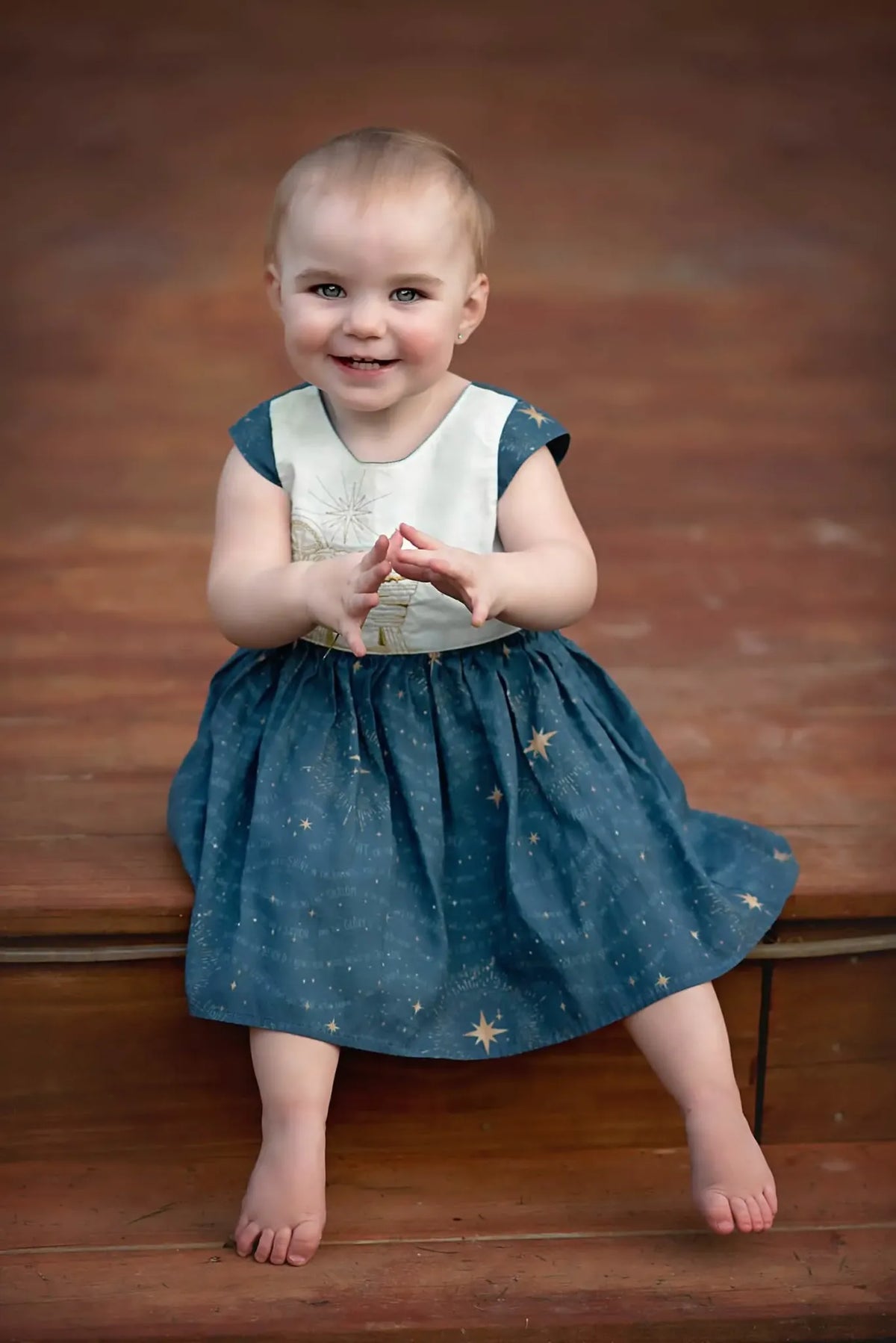 A smiling baby sits wearing a blue dress with gold stars and a white top.