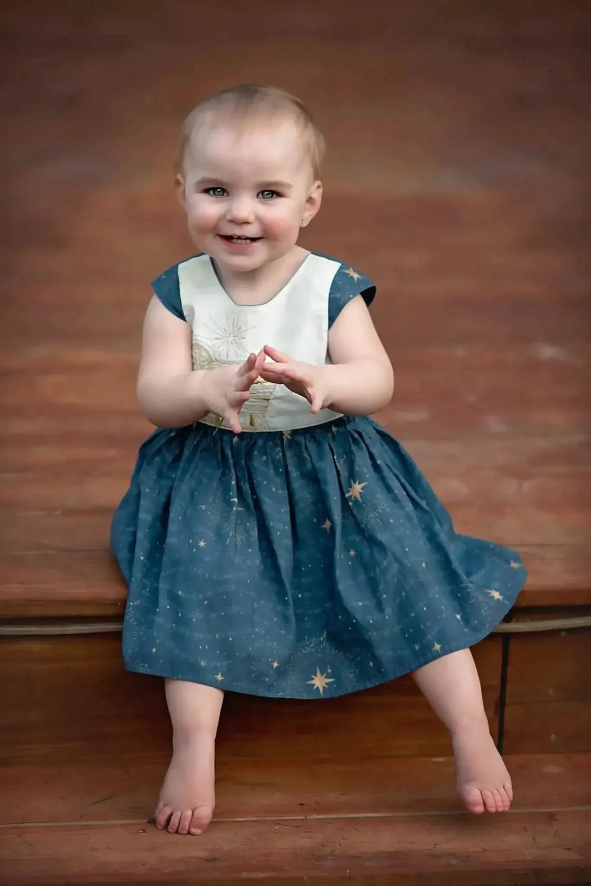 A smiling baby girl sits on wooden steps wearing a celestial blue dress with gold star print.