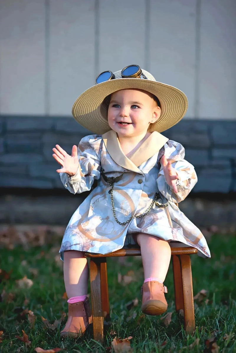 Toddler girl in gold clock-patterned faith dress with hat and goggles on stool.