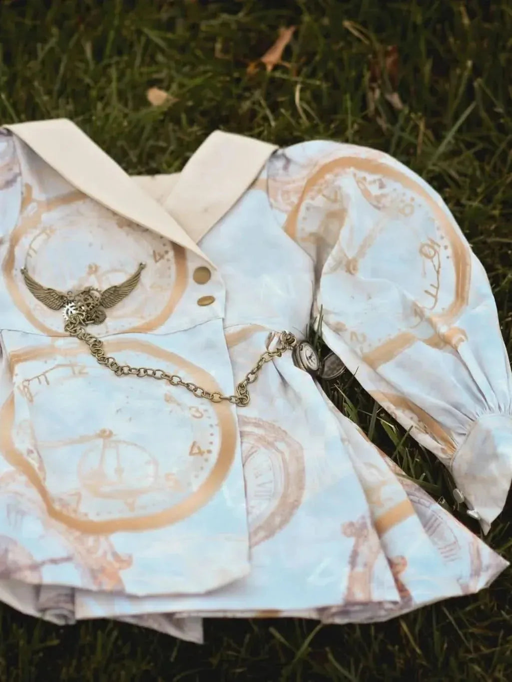 A light blue dress with gold clock designs and a pocket watch accessory lies on grass.