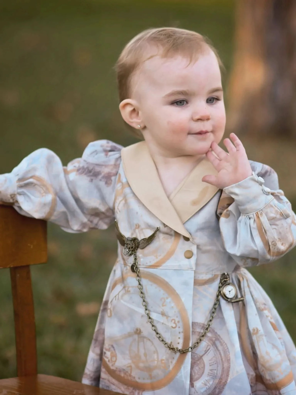 A baby in a dress with a clock and map pattern leans on a wooden chair.