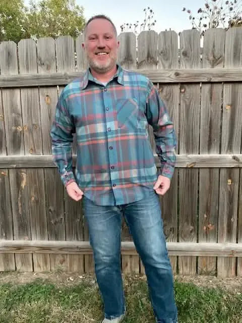 A man smiles wearing a blue, red, and tan plaid button-up shirt and jeans.
