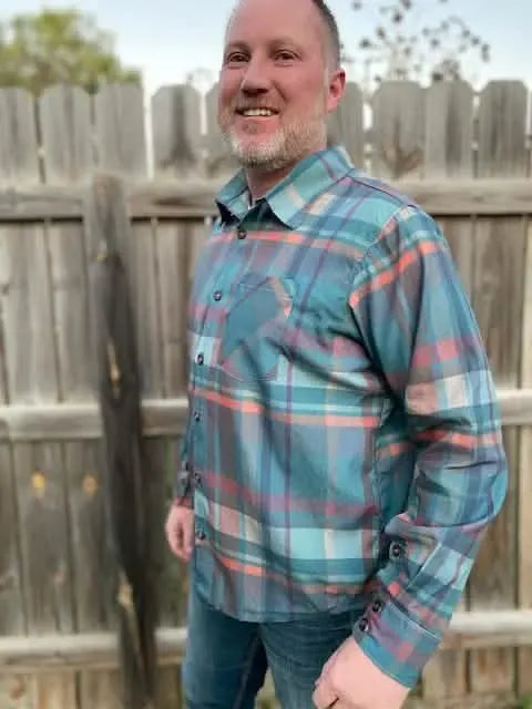 A man smiles wearing a blue, orange, and grey plaid long-sleeved shirt.
