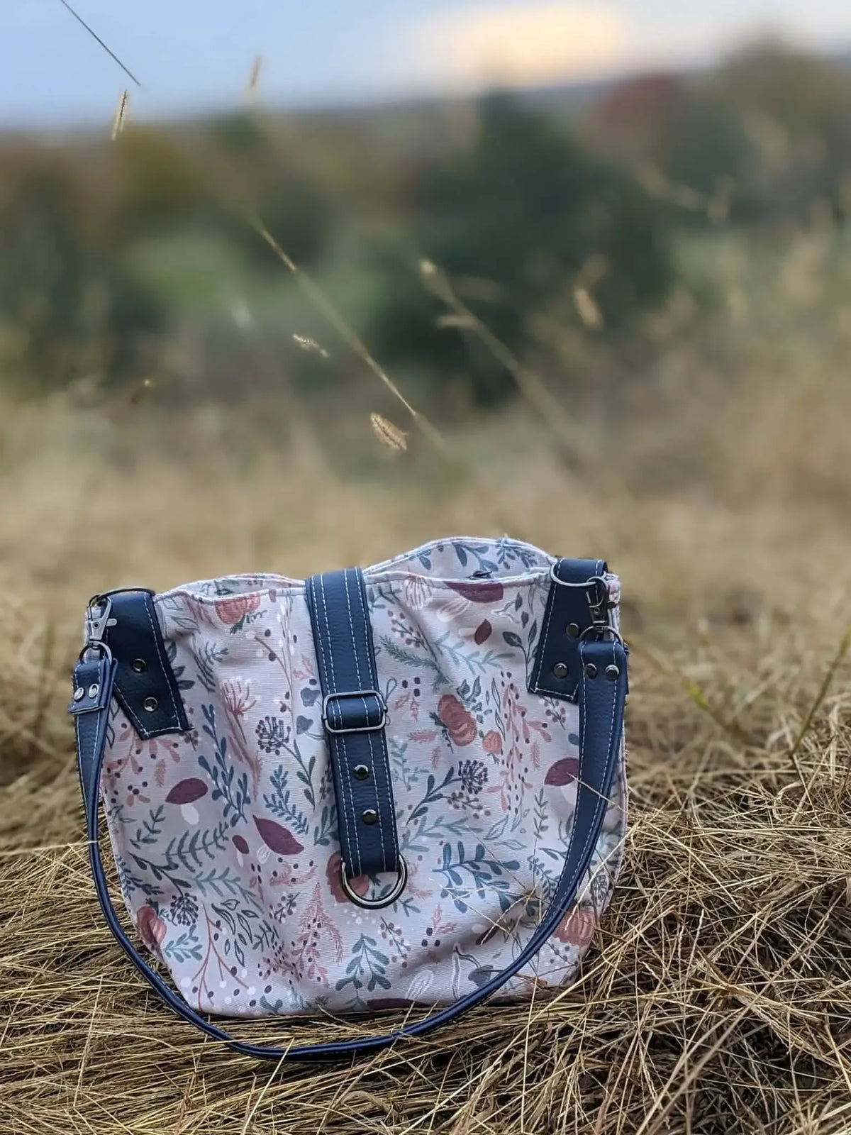 A tan bag with a forest foliage pattern and dark blue straps sits in dry grass.