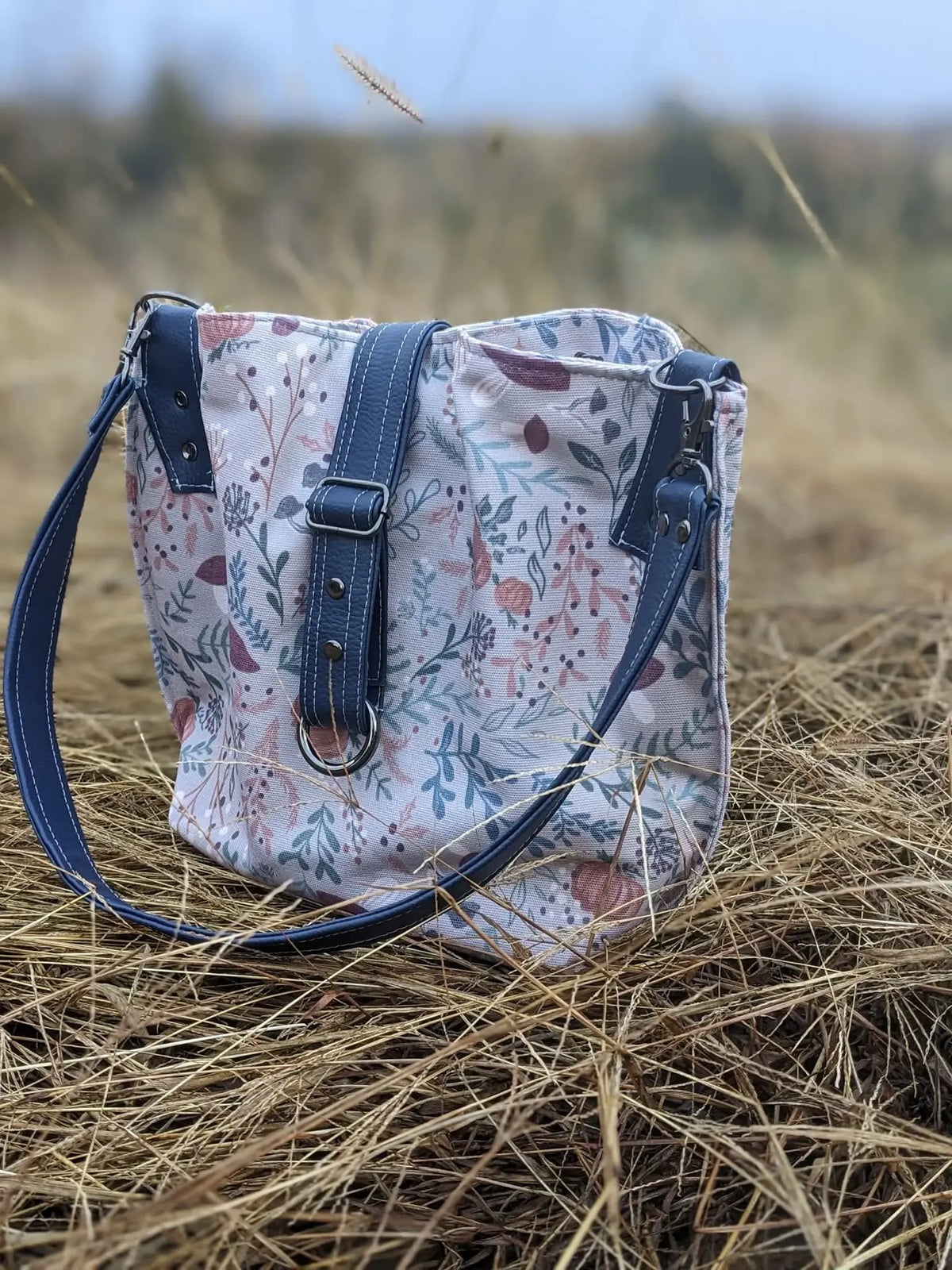 A tan bag with a forest foliage pattern and dark blue straps sits on dry grass.