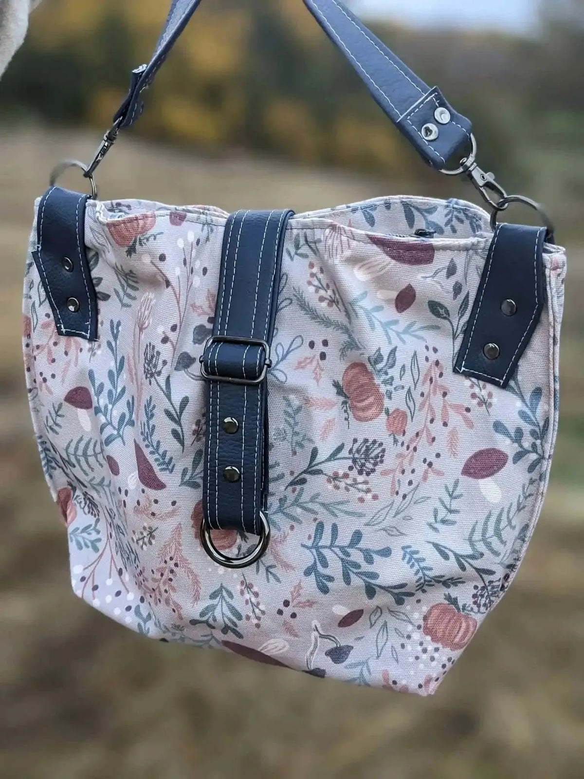 A tan botanical fabric bag with a forest foliage pattern and dark blue straps.