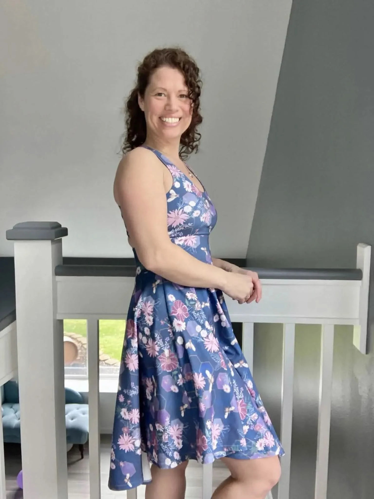 A woman smiles, wearing a blue floral dress with purple flowers and bees.