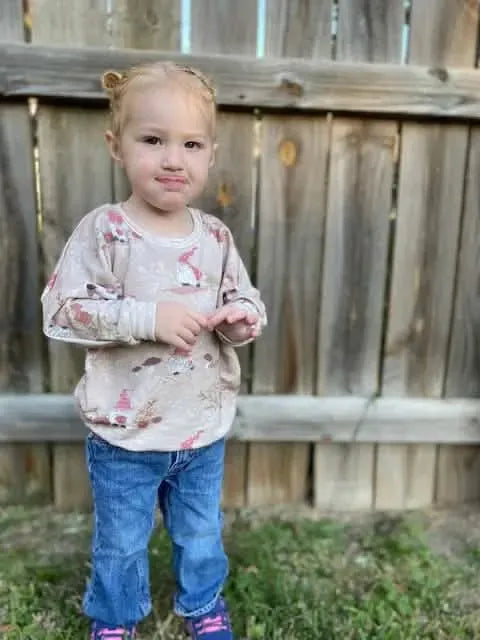 A young child wears a tan shirt with a gnome toile pattern and blue jeans.