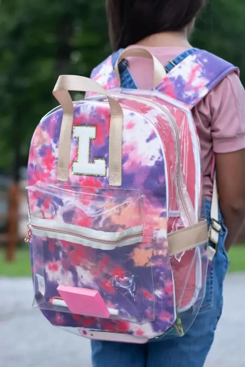 A person wears a tie-dye backpack with a clear front pocket and an "L" patch.
