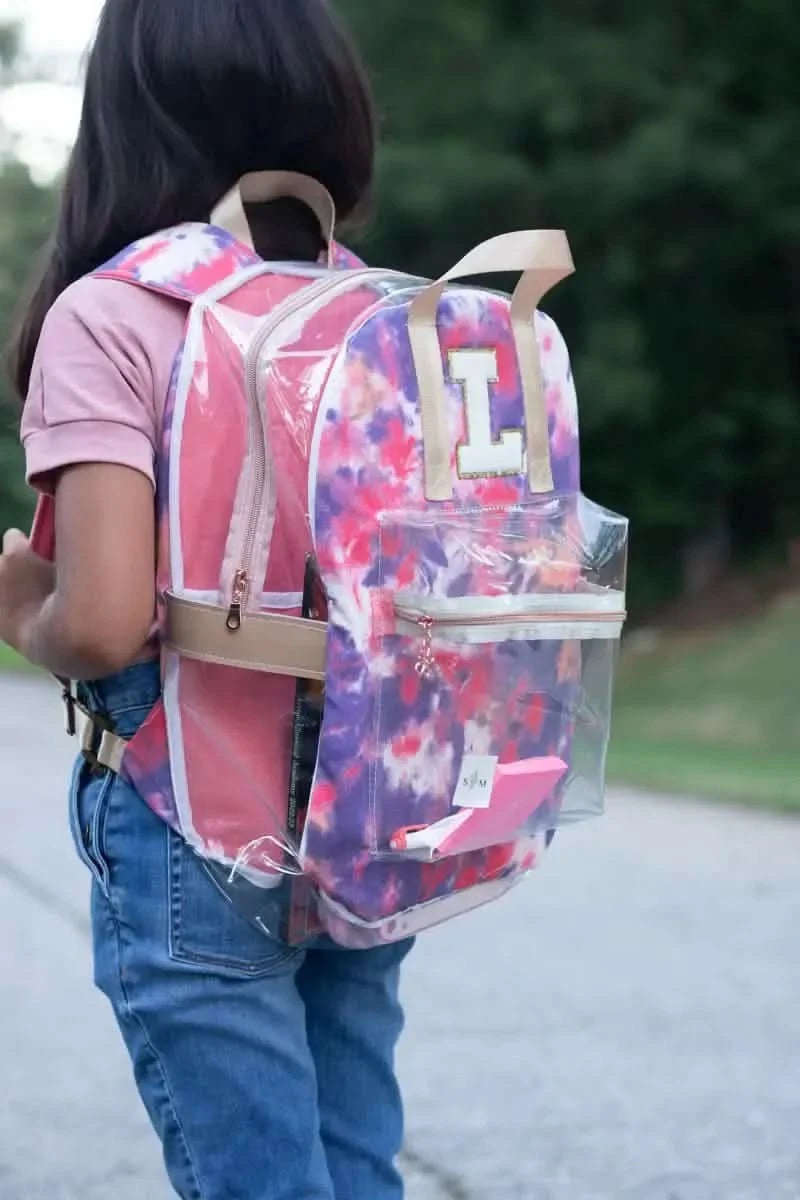 A child wears a clear backpack with a purple and pink tie-dye pattern and a white "L" patch.