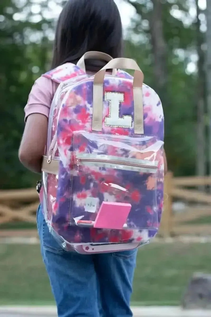 A person wears a clear backpack with a purple and pink tie-dye insert and a large "L" patch.