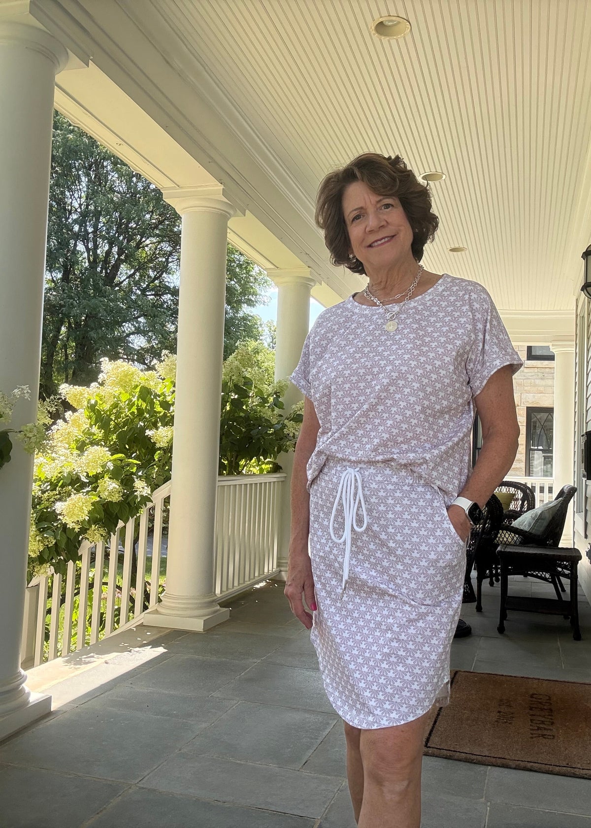A woman stands on a white-columned porch wearing a white short-sleeve dress with a small pink polka dot pattern and a matching tie belt.