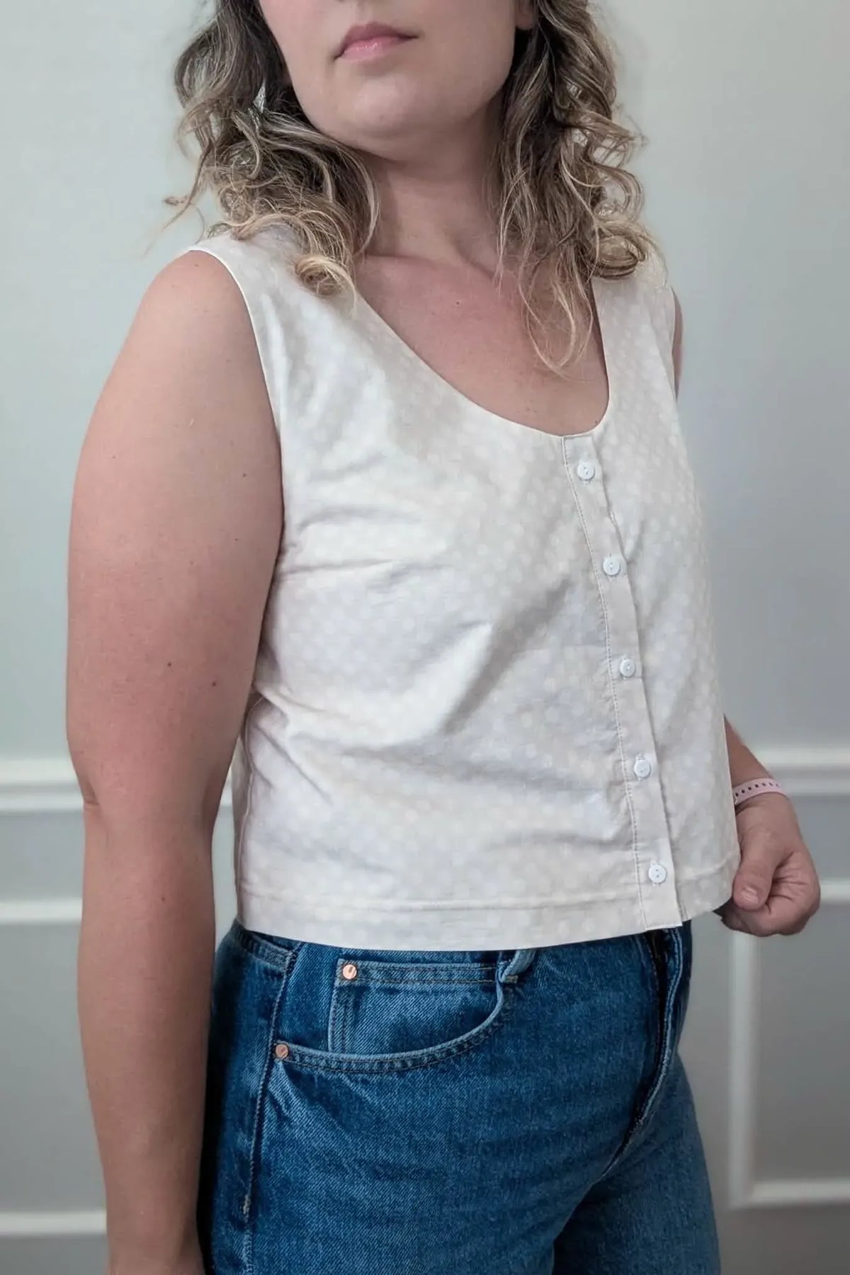 A woman wears a light-colored, polka-dotted sleeveless top with buttons and blue jeans.