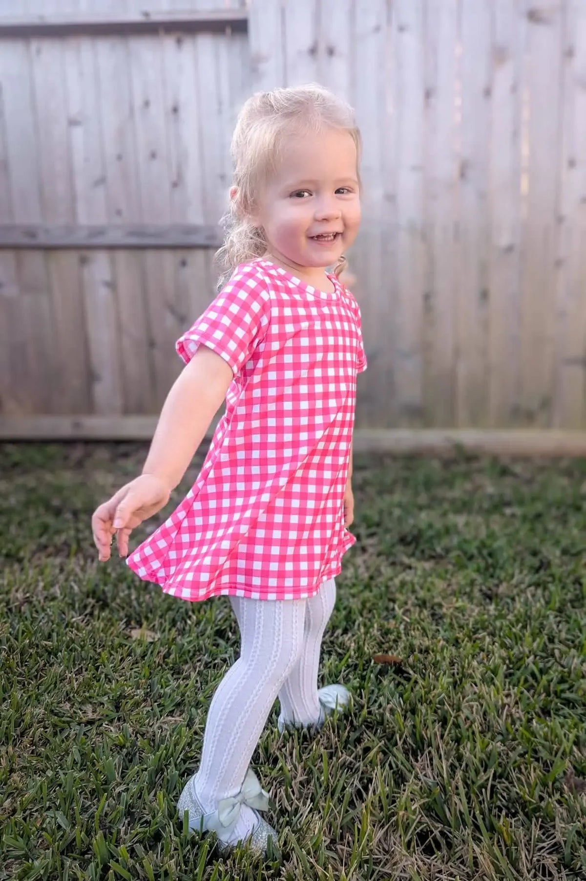 A young girl smiles while wearing a hot pink gingham dress and white tights.
