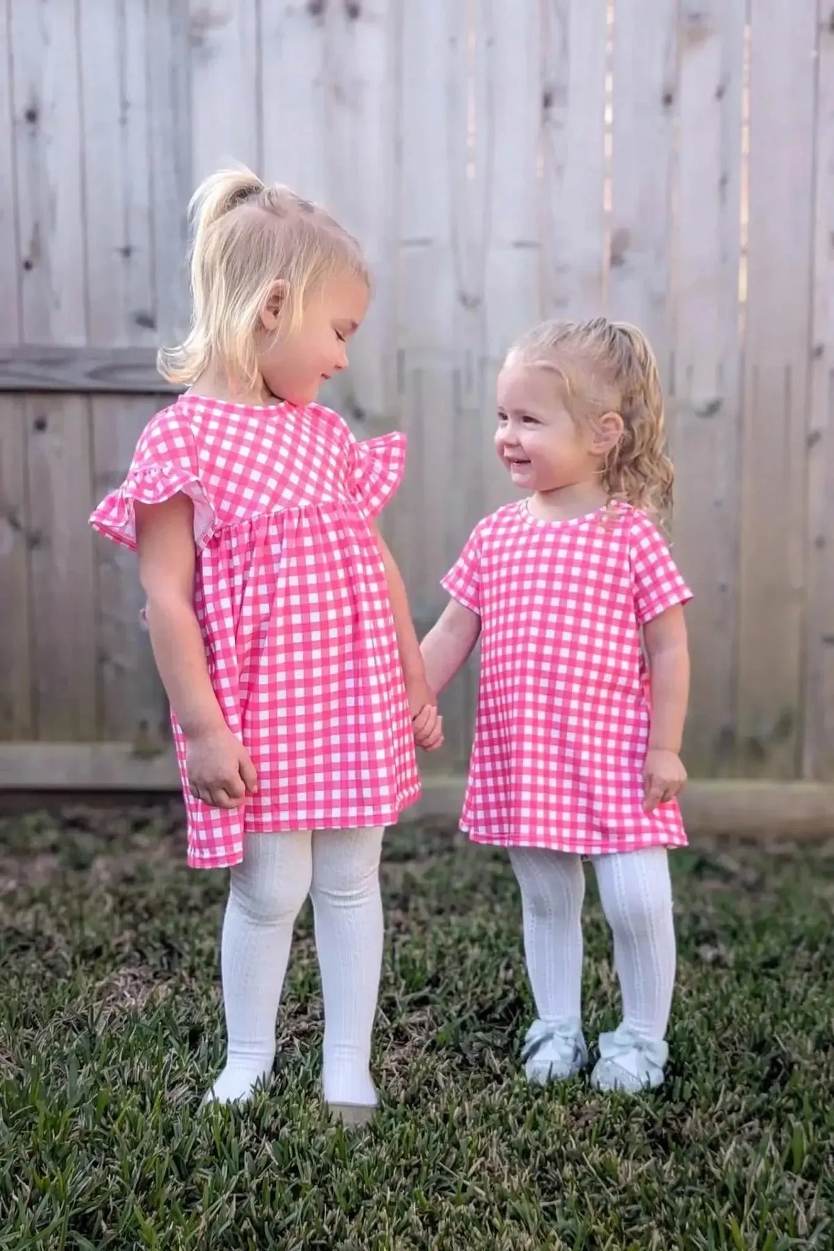 Two young girls in pink gingham dresses holding hands outdoors.