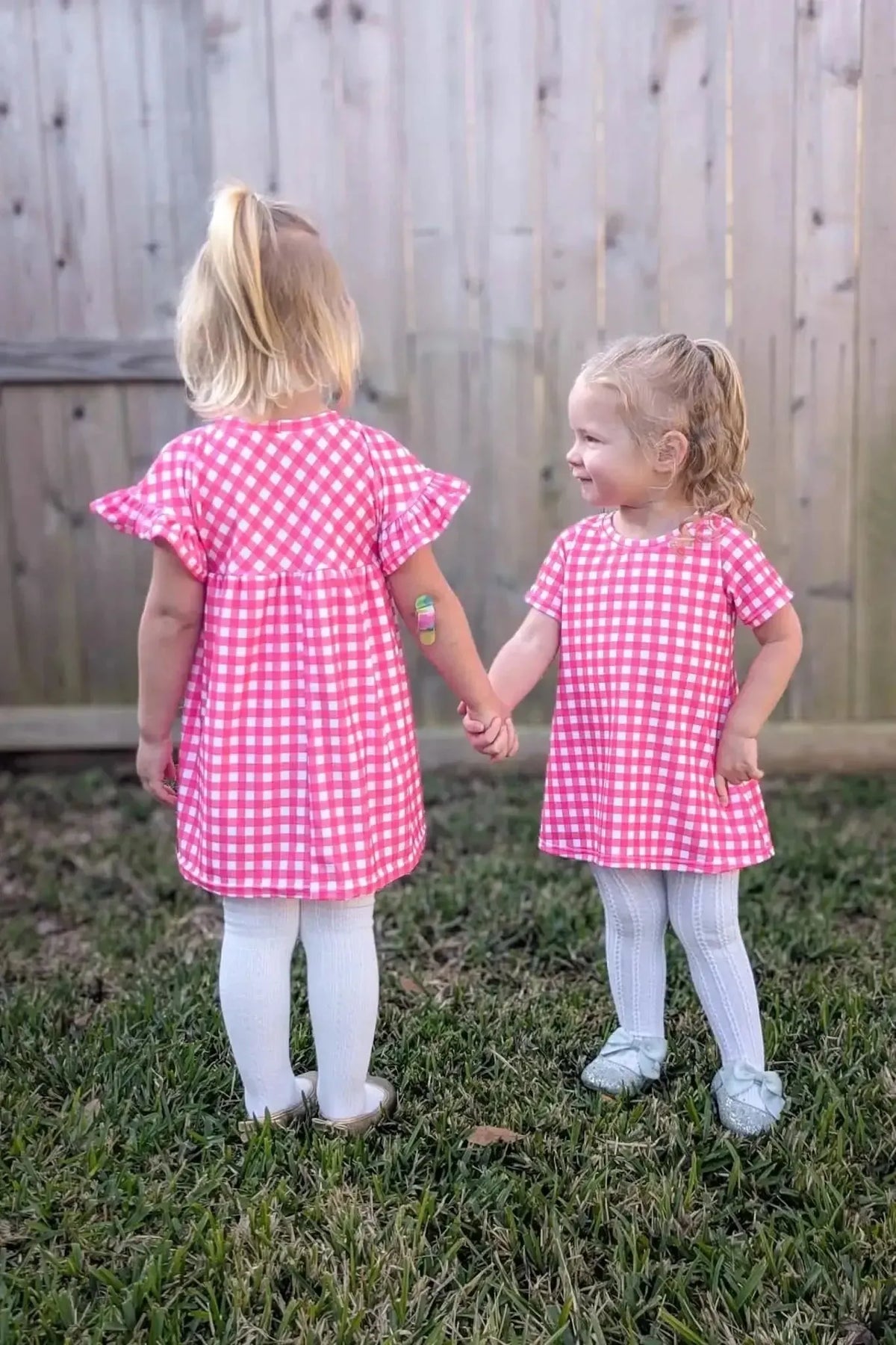 Two young girls in hot pink gingham dresses hold hands in a backyard.