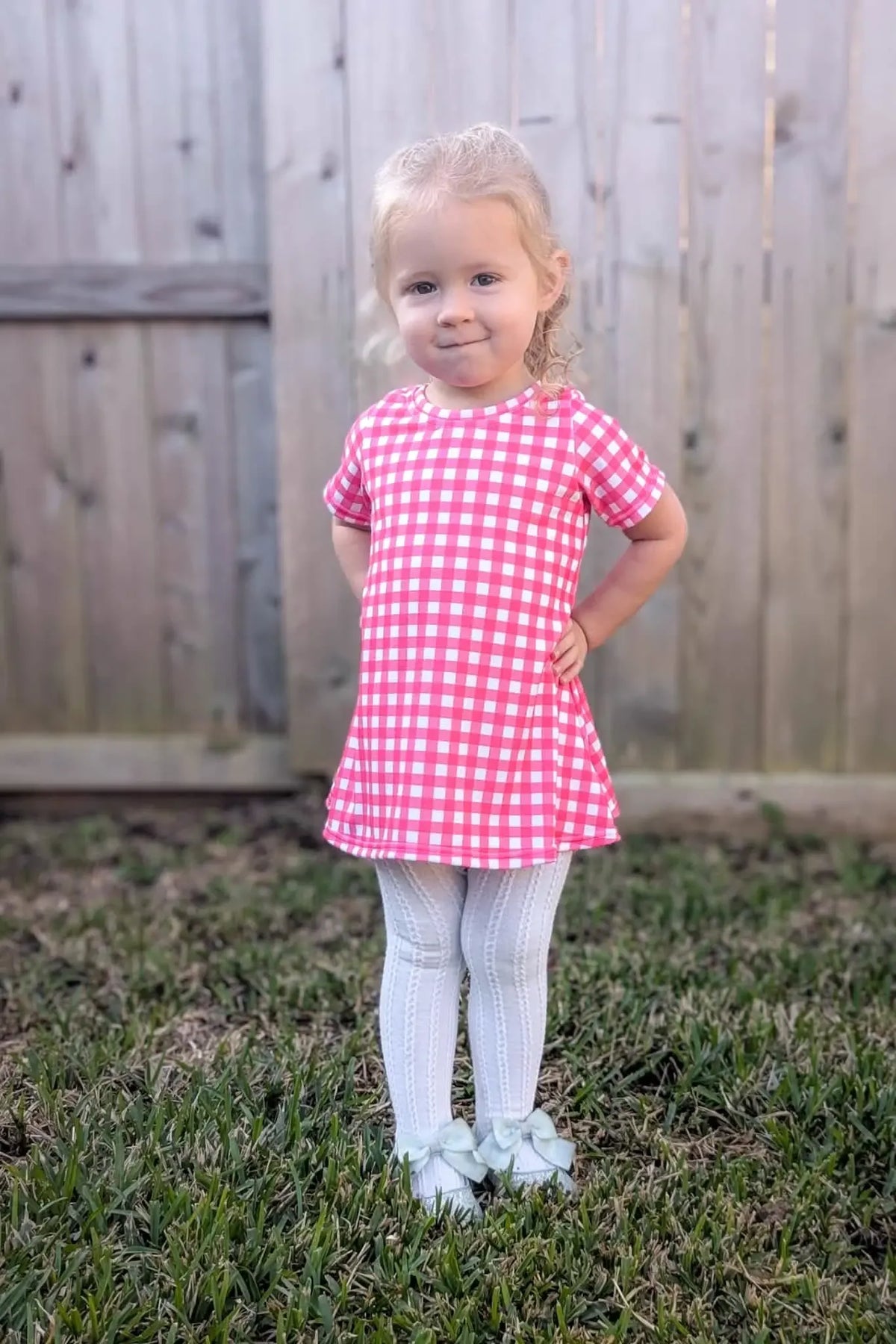 A young girl in a hot pink gingham dress and white tights stands in grass.