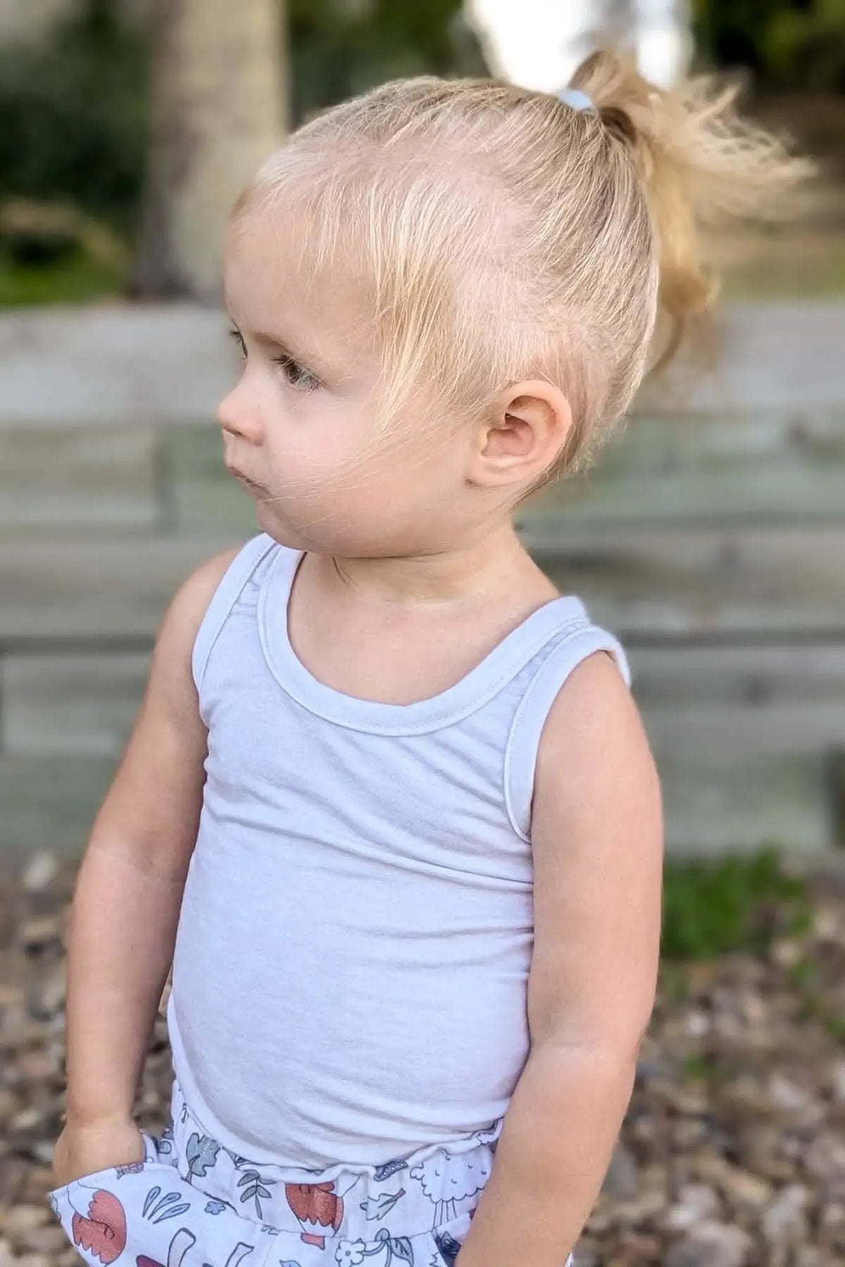 A young child with blonde hair in a ponytail wears a white tank top and patterned shorts.