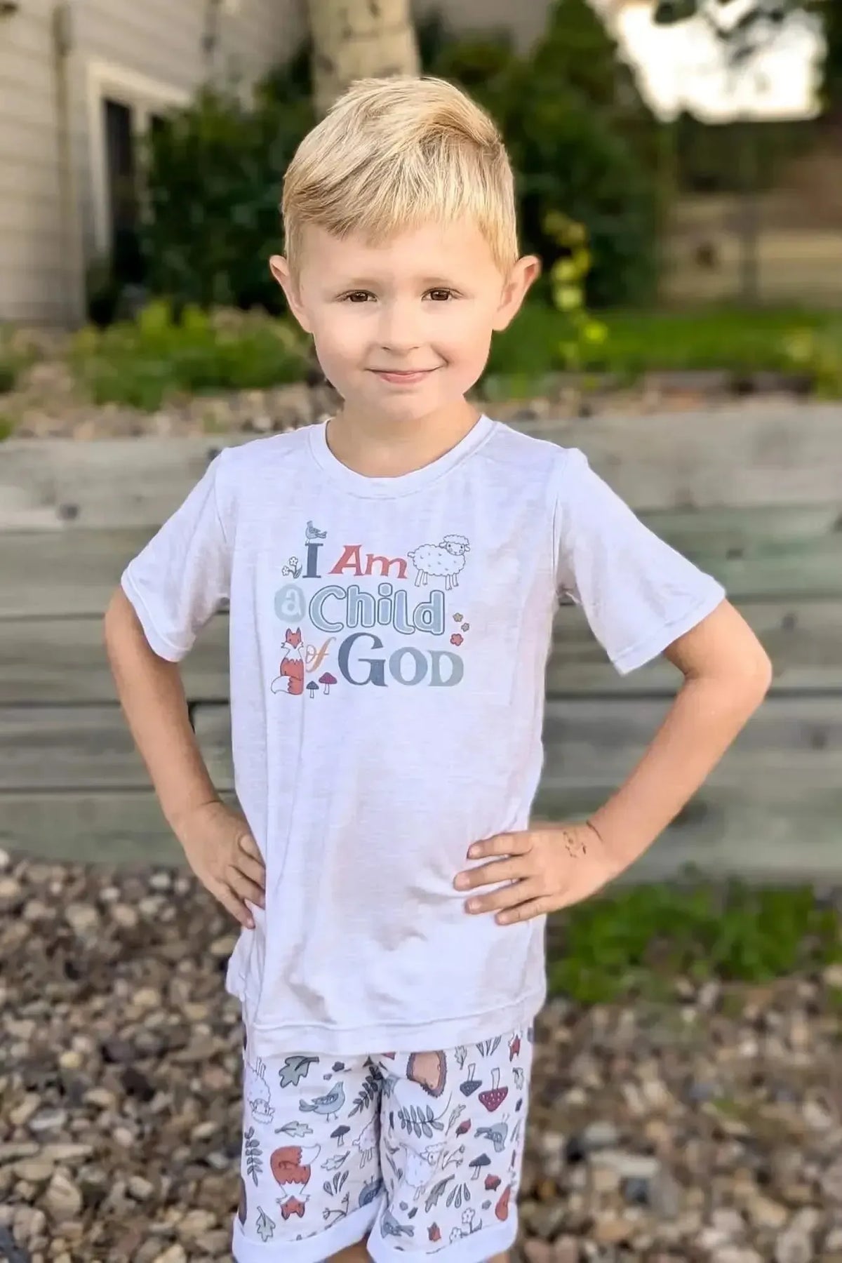 Boy in white "I Am Child of God" t-shirt and patterned shorts stands outdoors.