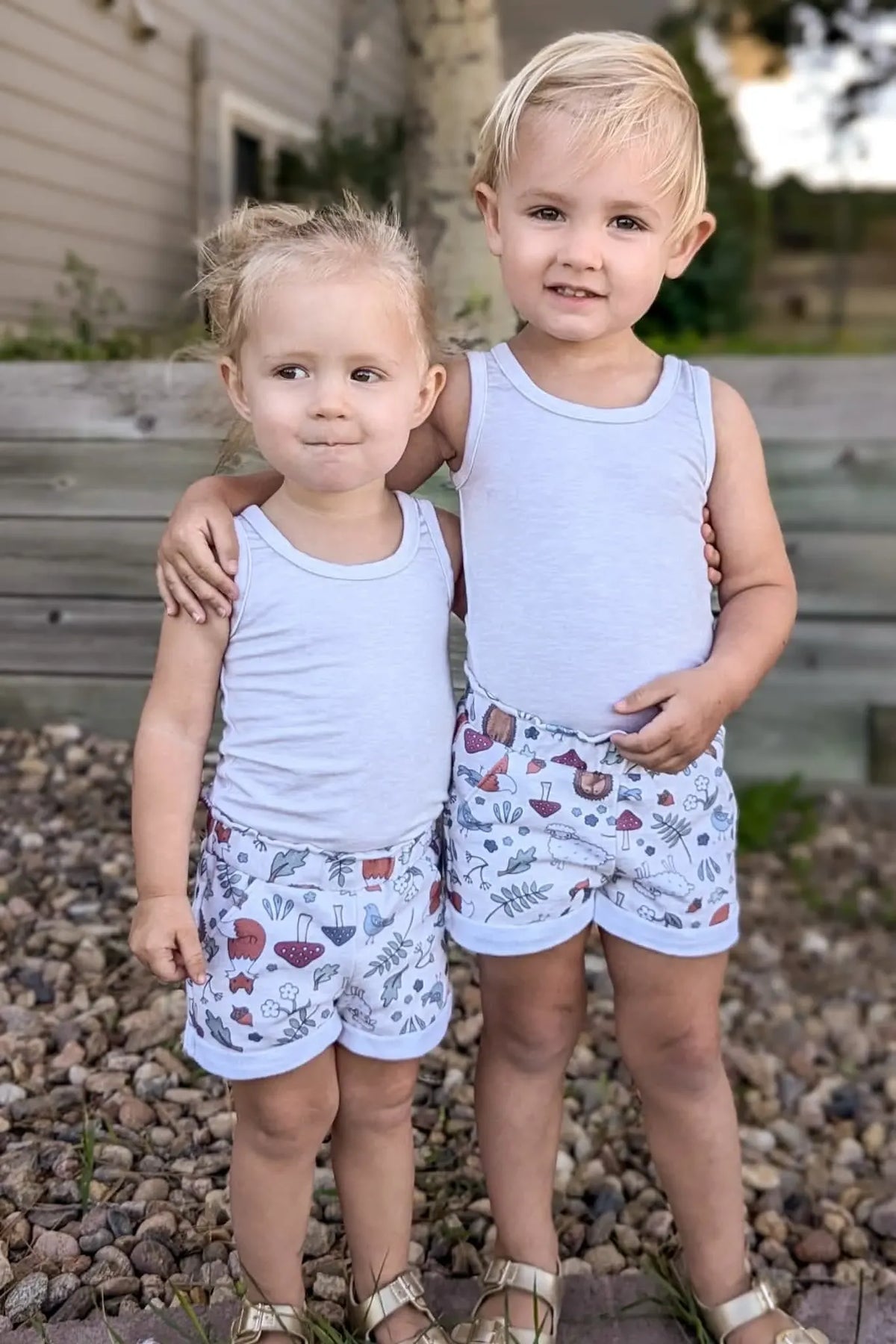 Two children in white tank tops and patterned shorts stand on a rocky ground.