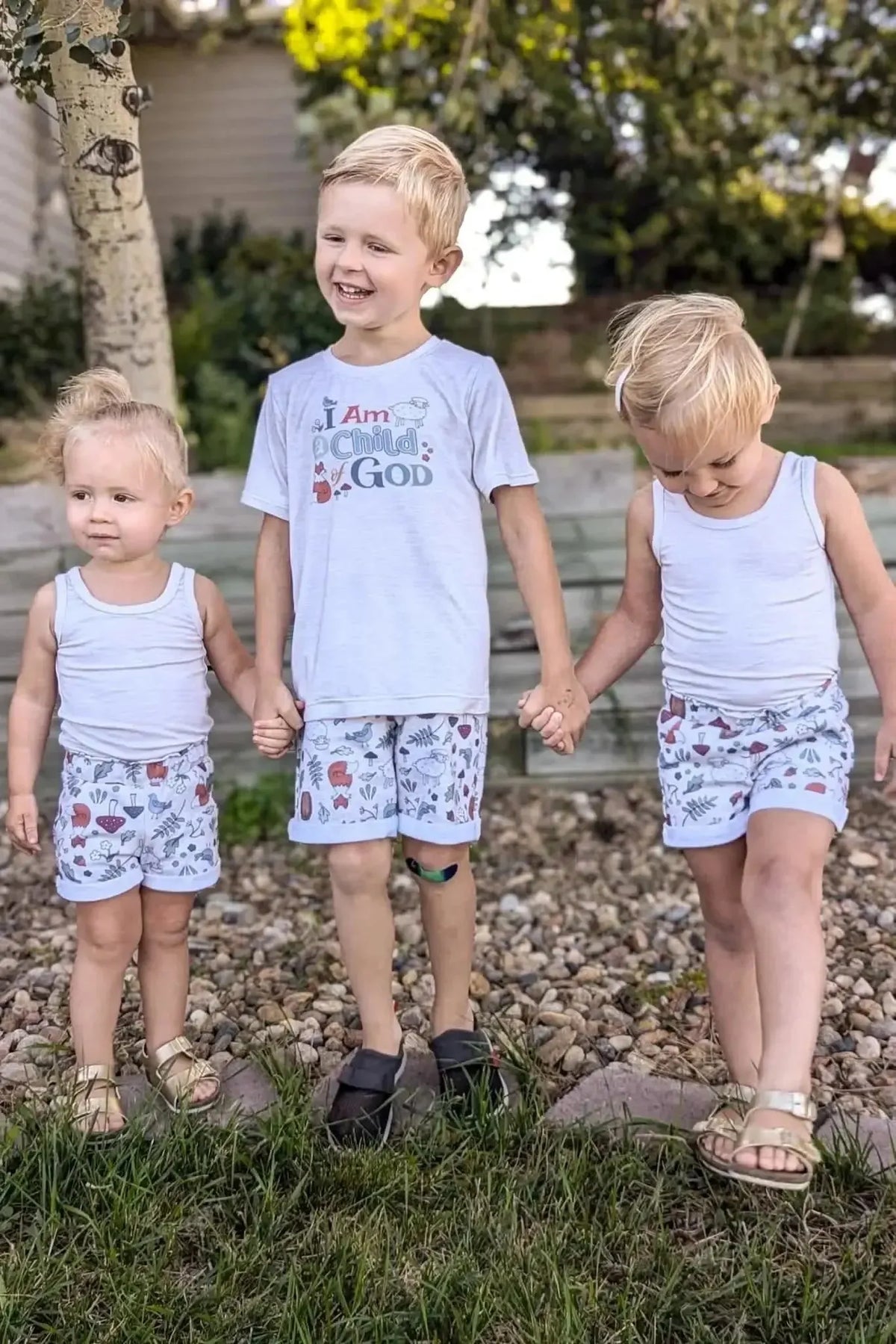 Three young children wearing white outfits with woodland creatures print on shorts, holding hands outdoors.