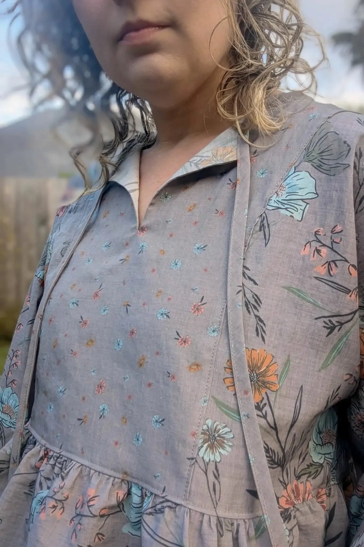 A woman wears a taupe floral dress with a collared neckline and ruffled hem.