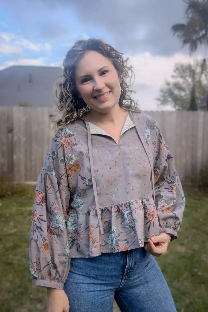 A woman smiles wearing a taupe floral blouse with blue jeans in a backyard.