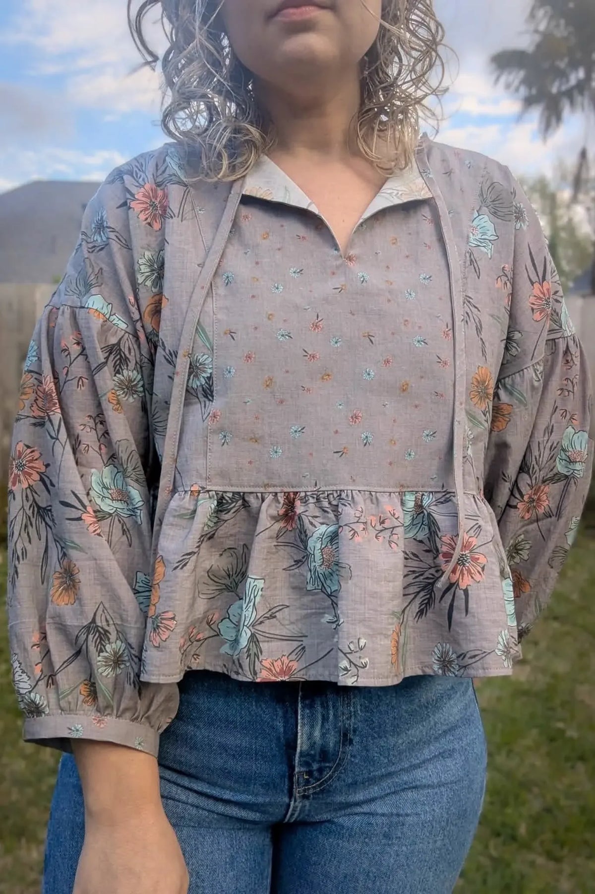 A person wears a taupe floral blouse with a ruffled hem and blue jeans.