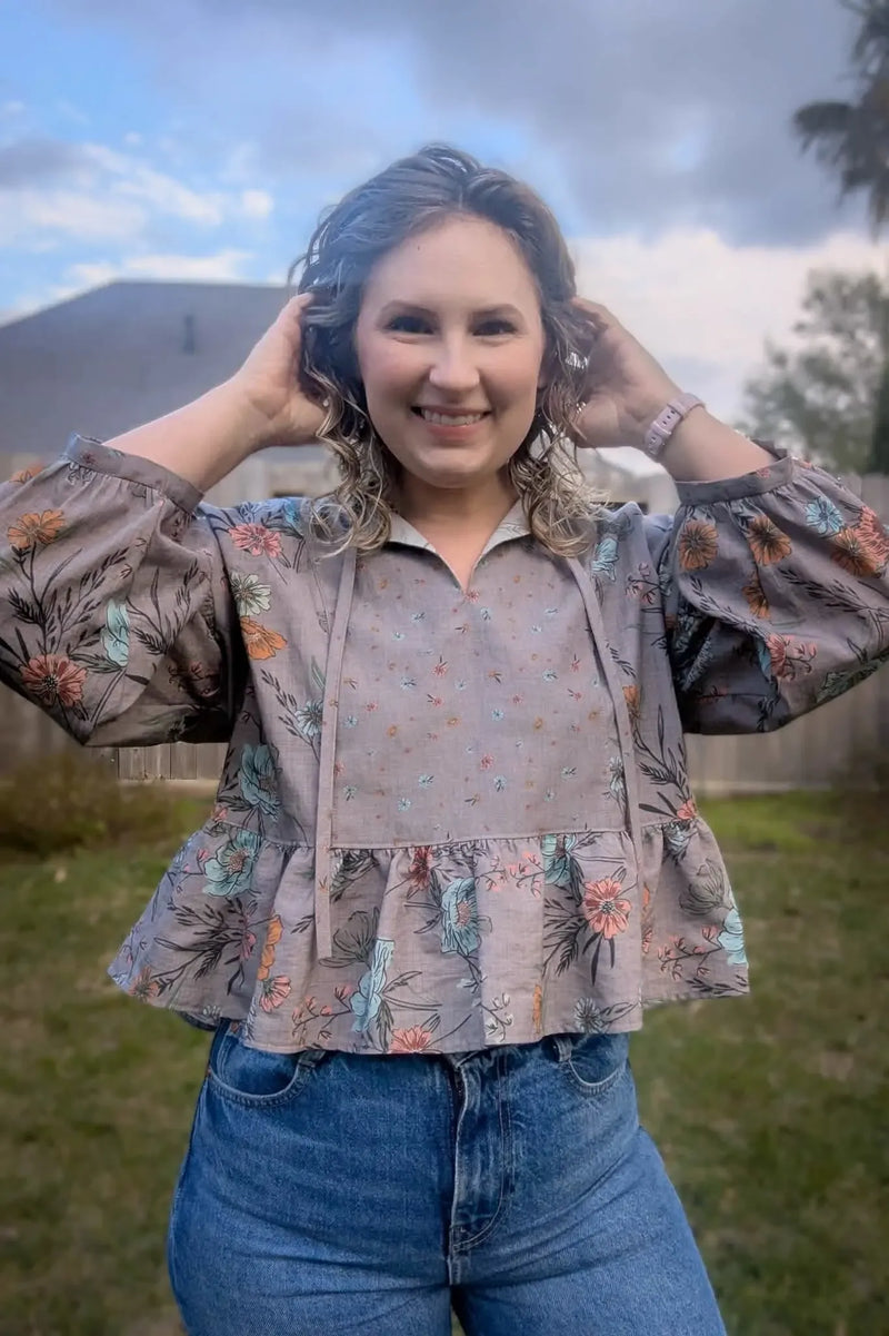 A woman smiles, wearing a taupe floral top with puffed sleeves and blue jeans.