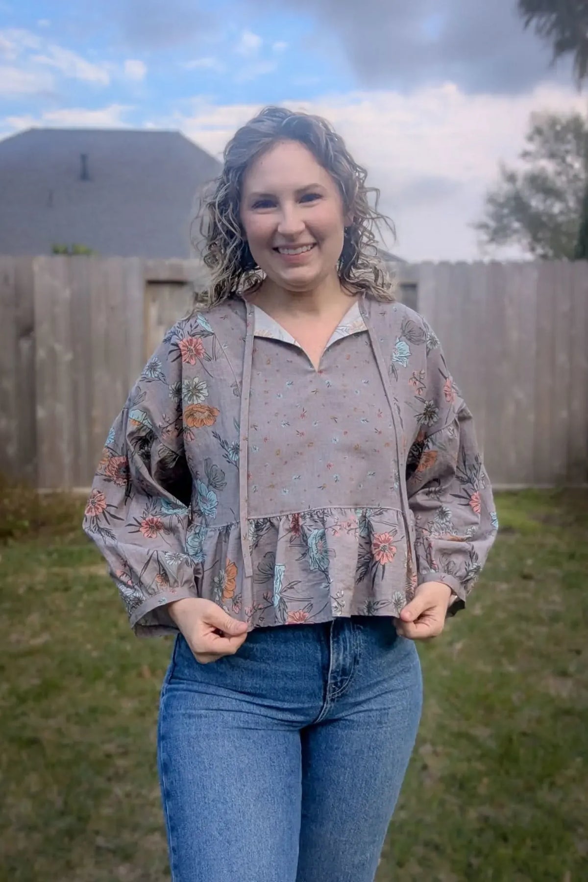 A woman wears a taupe floral print top with a ruffled hem and blue jeans.