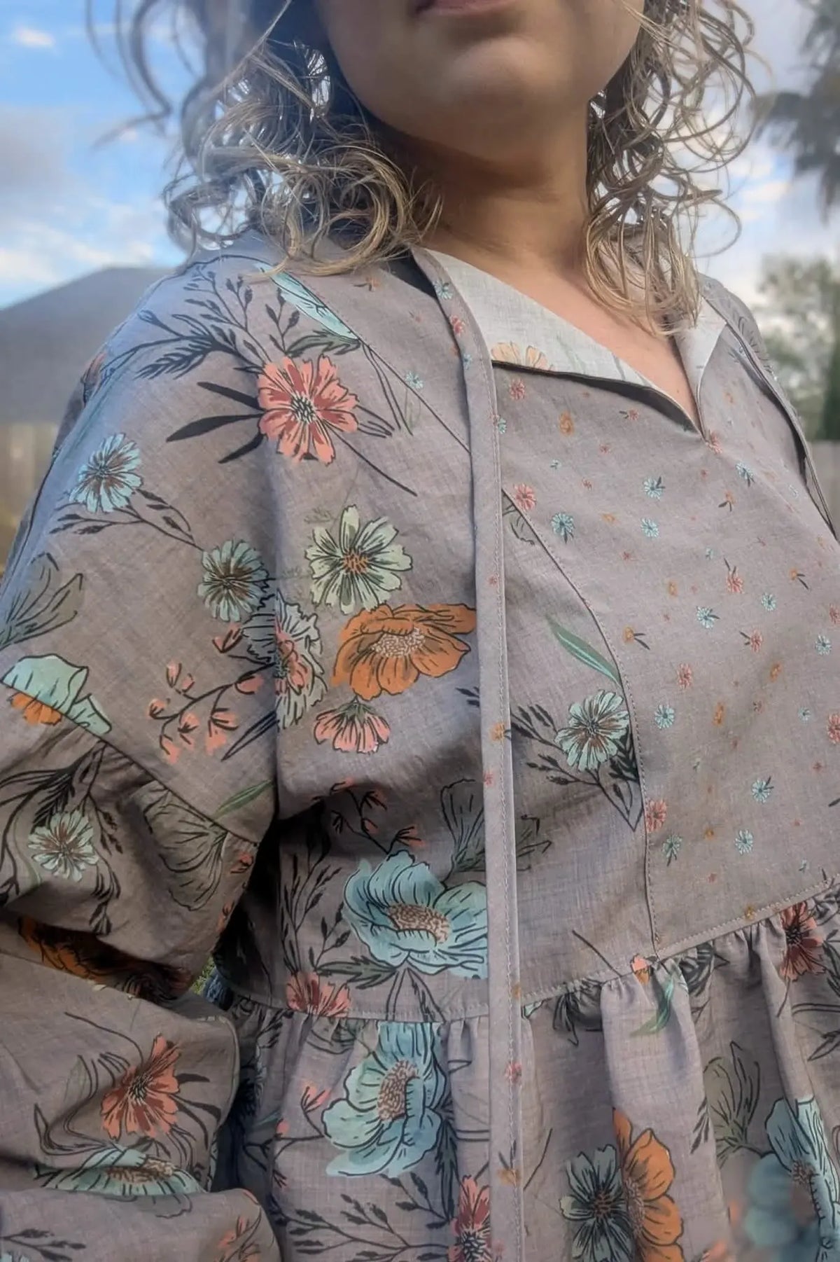 A person wears a taupe floral dress with a split neckline and ruffled hem.