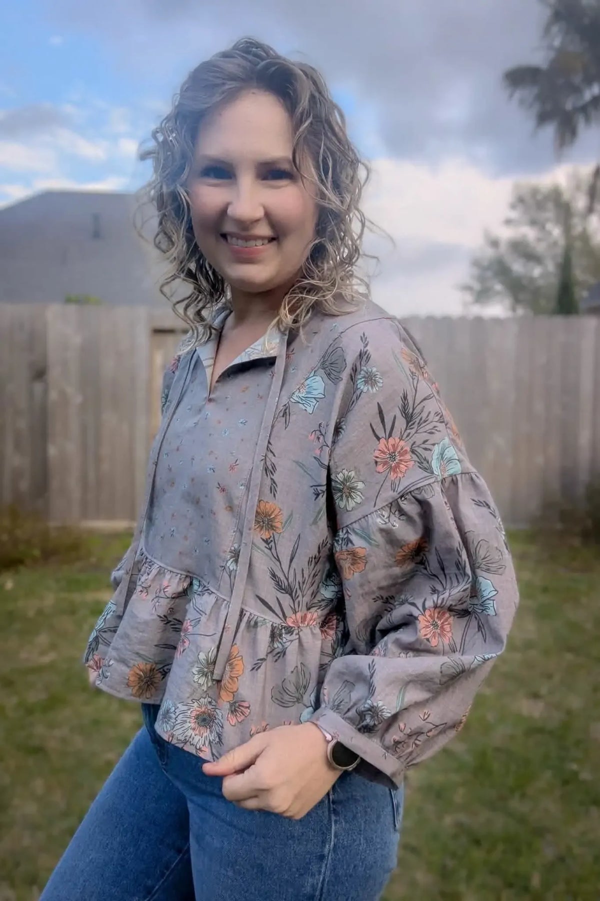 A woman smiles wearing a taupe floral top and blue jeans outdoors.