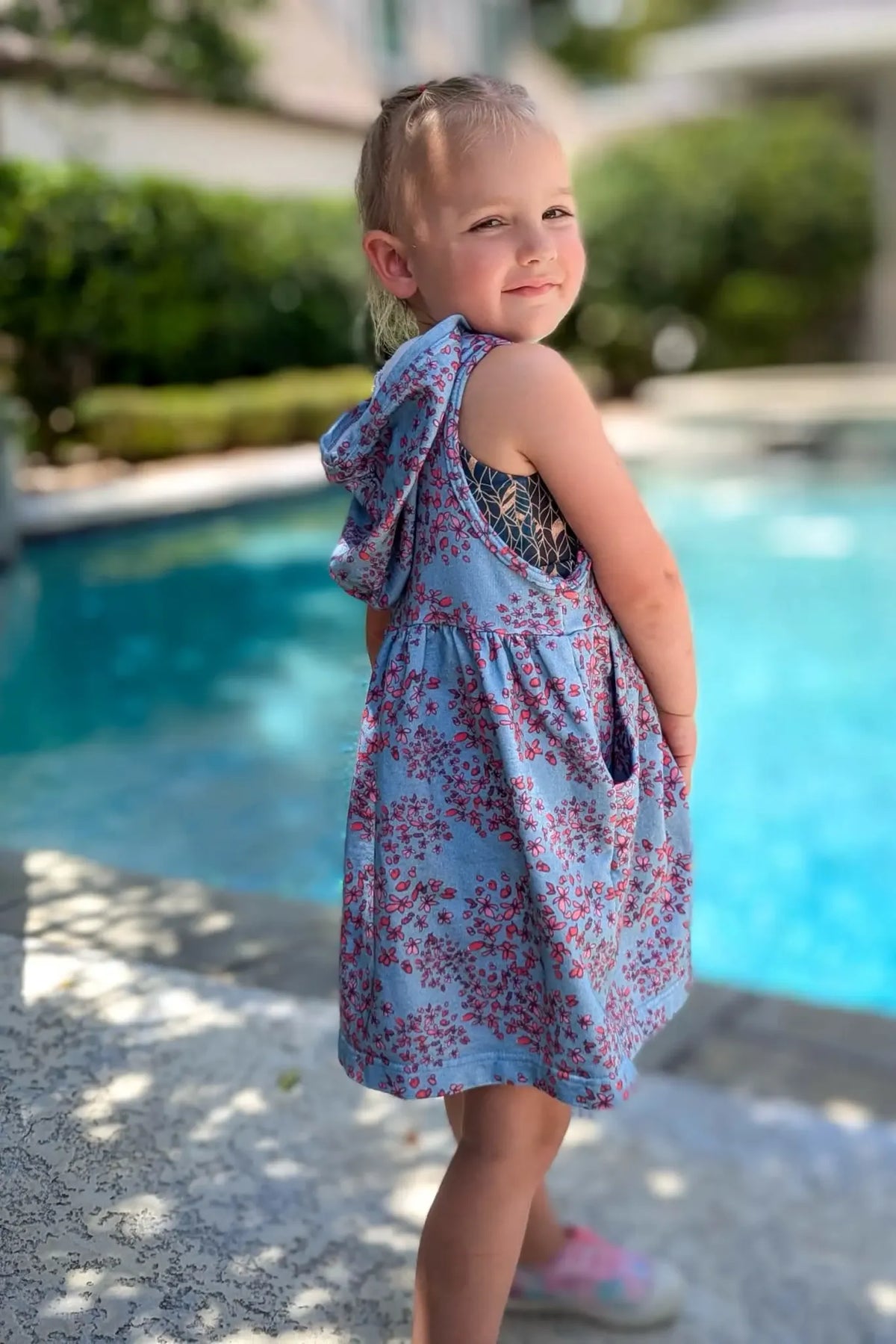 A young girl in a blue floral hooded dress stands by a pool.