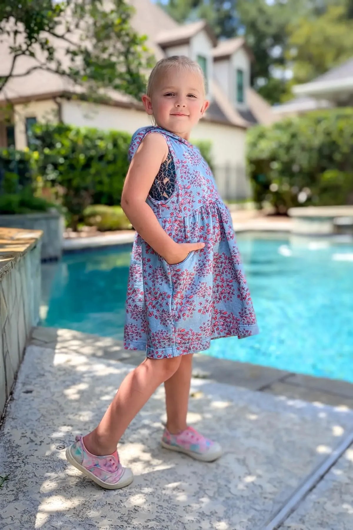 A young girl in a blue floral dress with pockets stands by a pool.