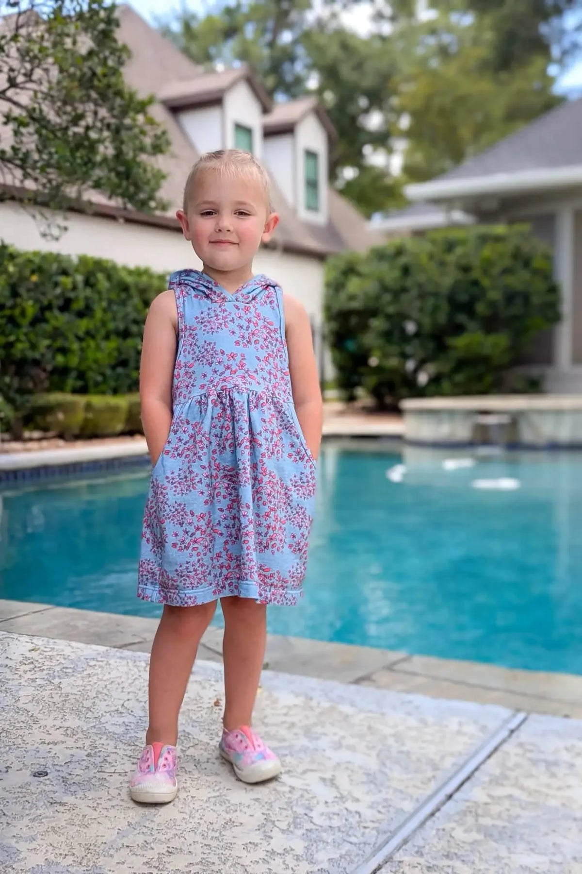 A young girl in a blue floral hooded dress stands by a pool.