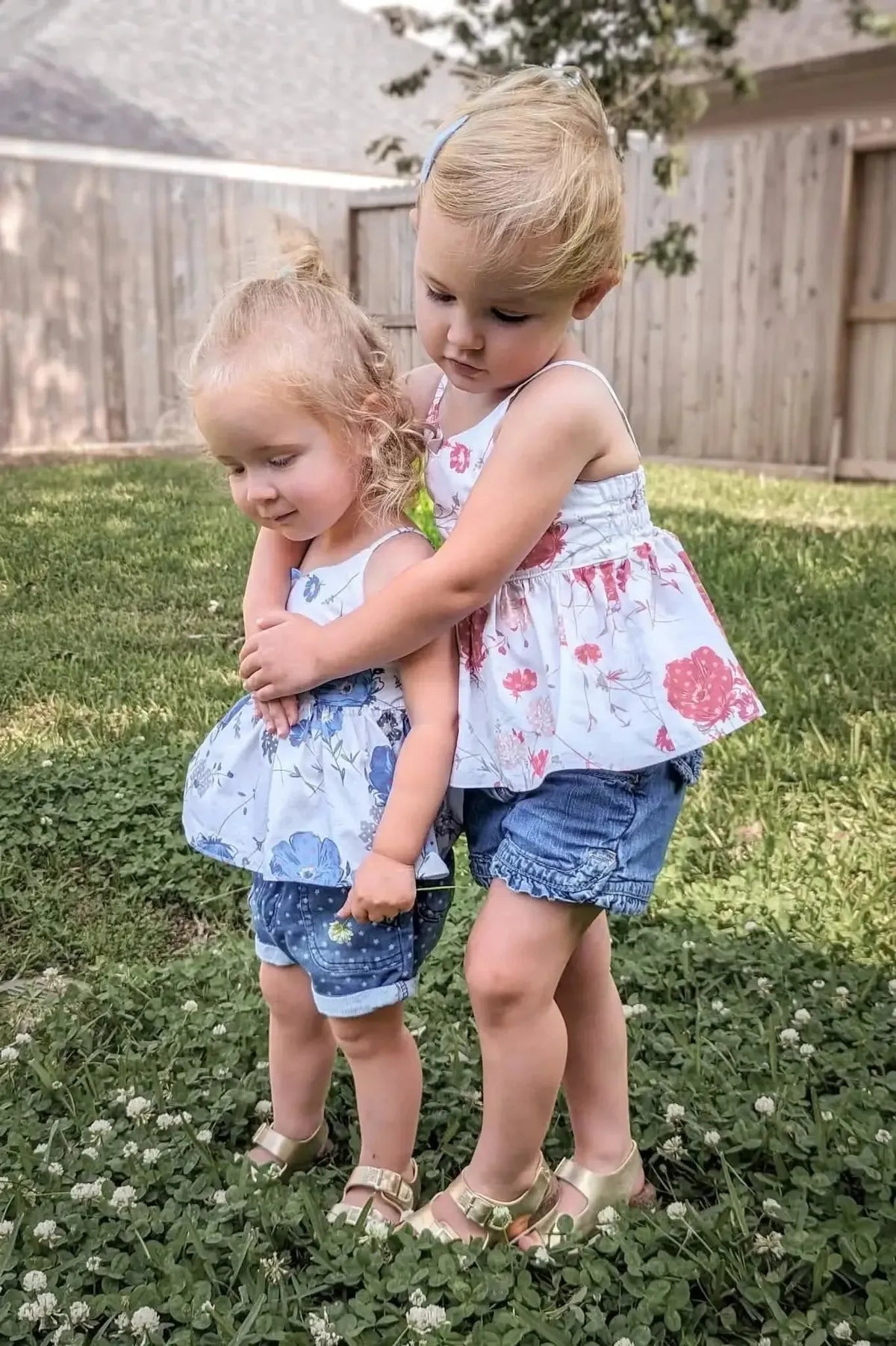 Two blonde toddler girls in floral dresses hugging on grass, showcasing red blooms print fabric.