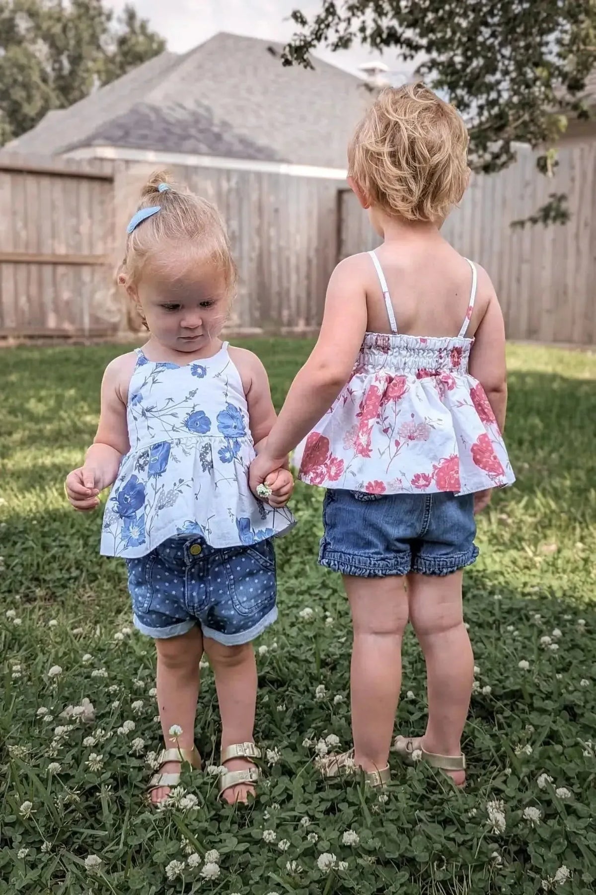Two toddlers wear dresses made from blue floral print fabric in a backyard.