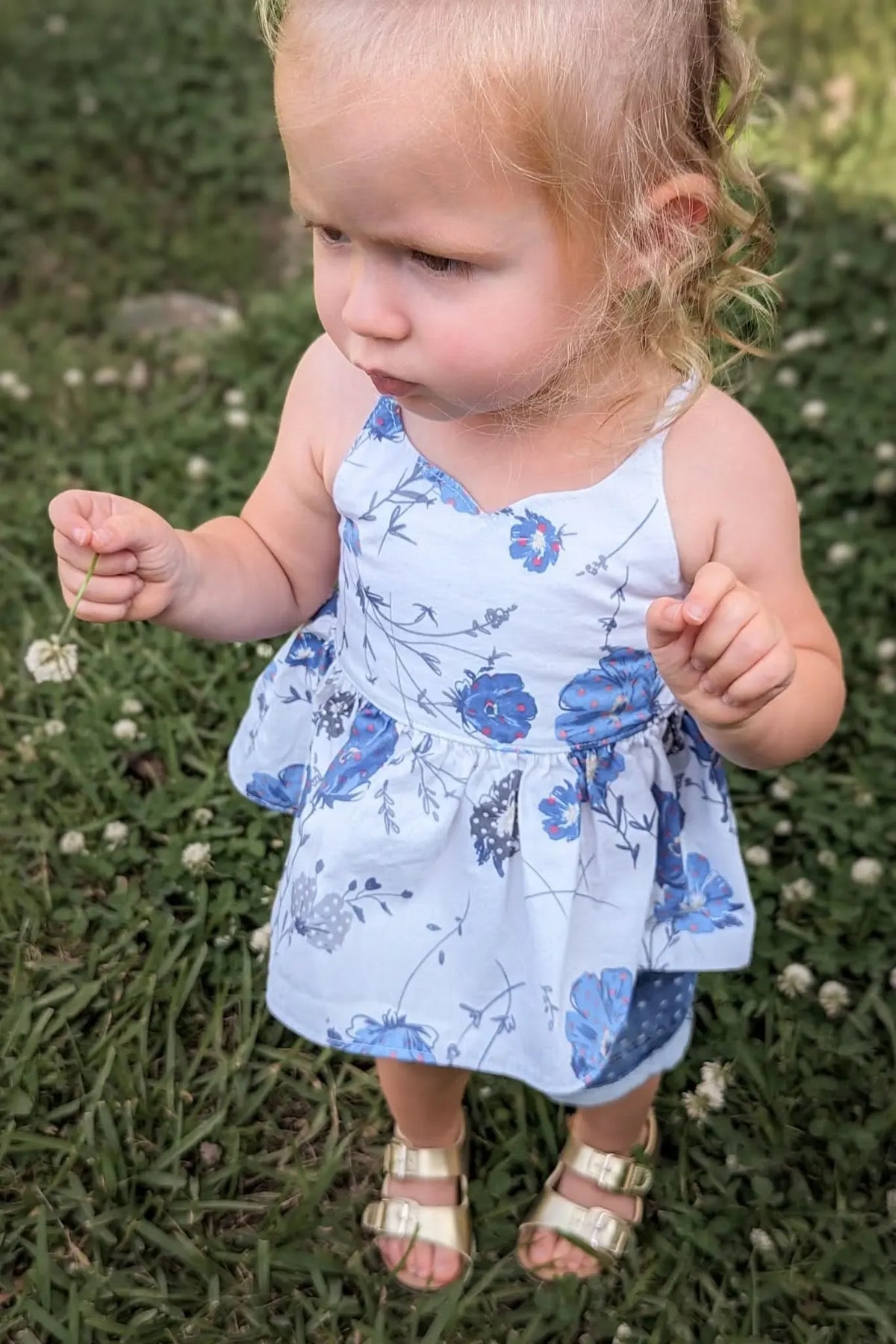 A young child in a blue floral dress and gold sandals holds a dandelion in grass.