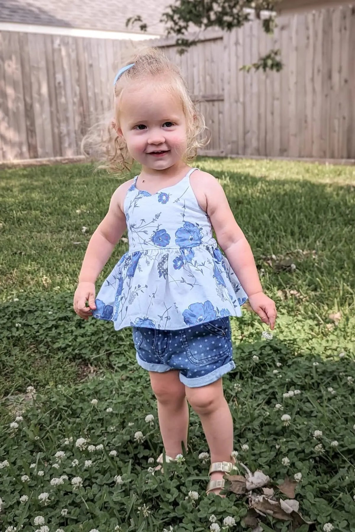 A young child in a blue floral top and denim shorts stands in a grassy yard.