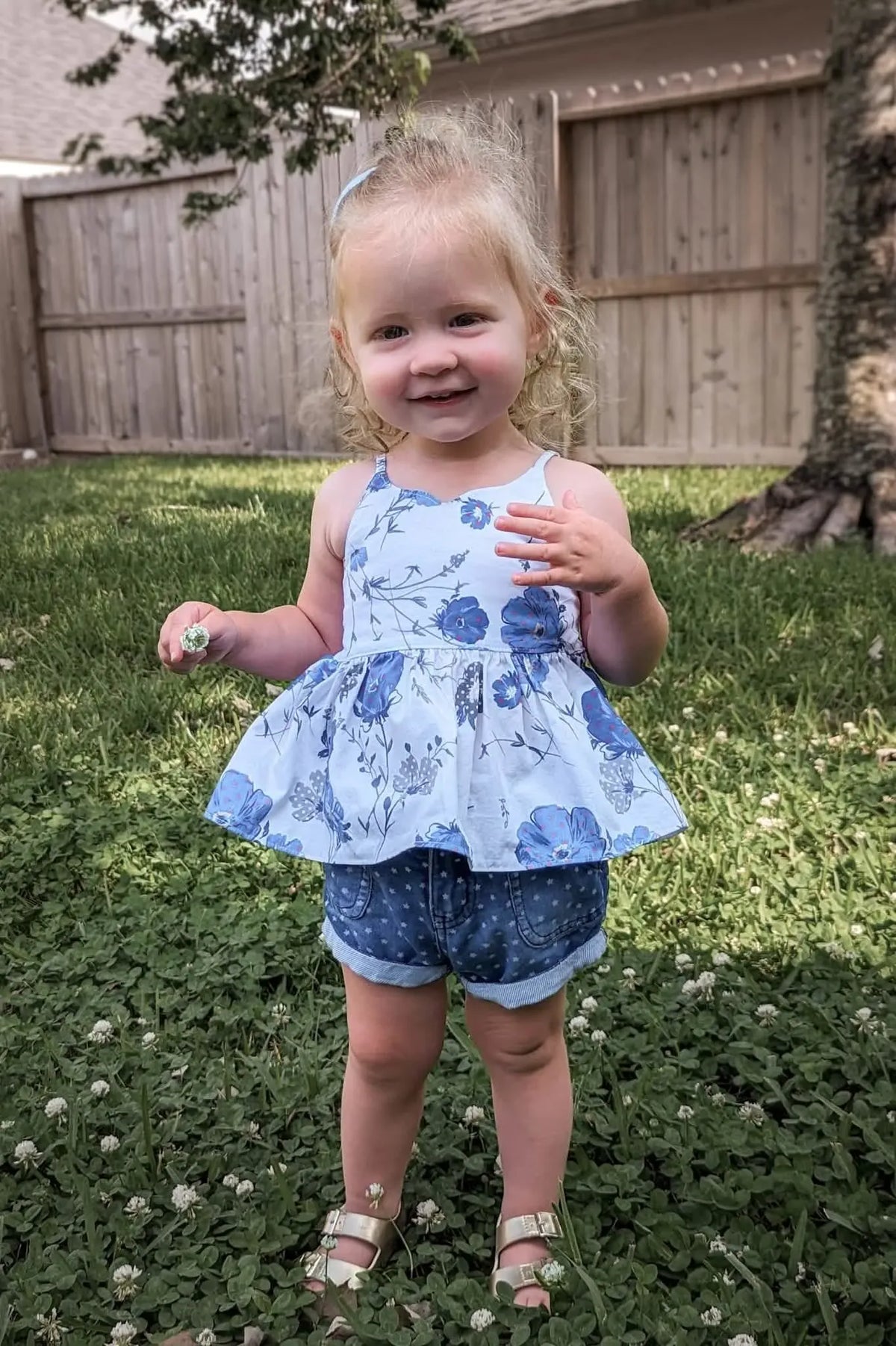 A young girl in a blue floral top and denim shorts stands in a grassy yard.