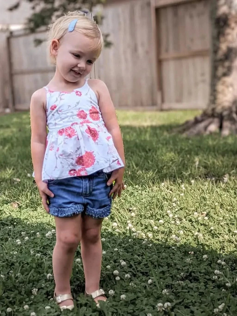 A young girl wears a white top with red floral print and denim shorts.