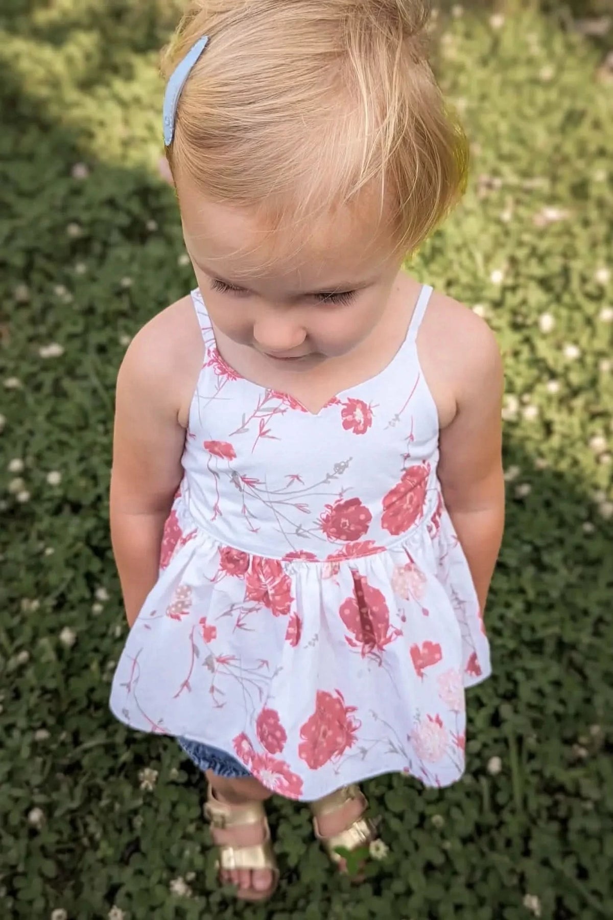 A young child in a white and red floral dress stands on green grass.