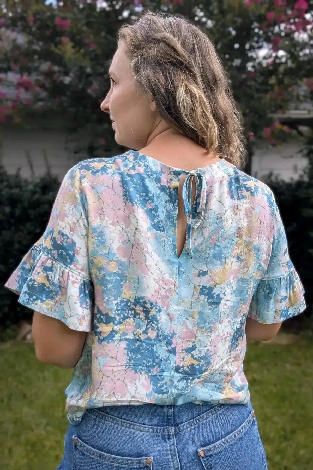 Back view of a woman wearing a blue-pink abstract floral print blouse with ruffled sleeves.