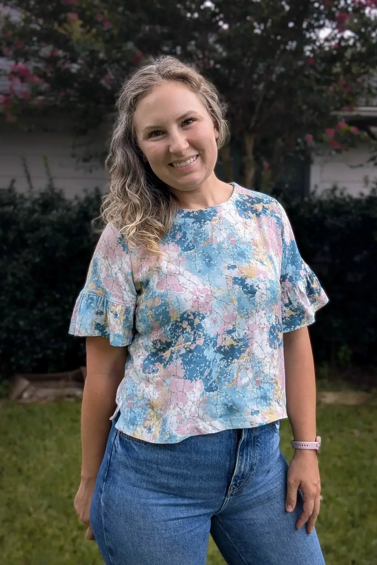 A woman wears a colorful, abstract-patterned top with ruffled sleeves and blue jeans.