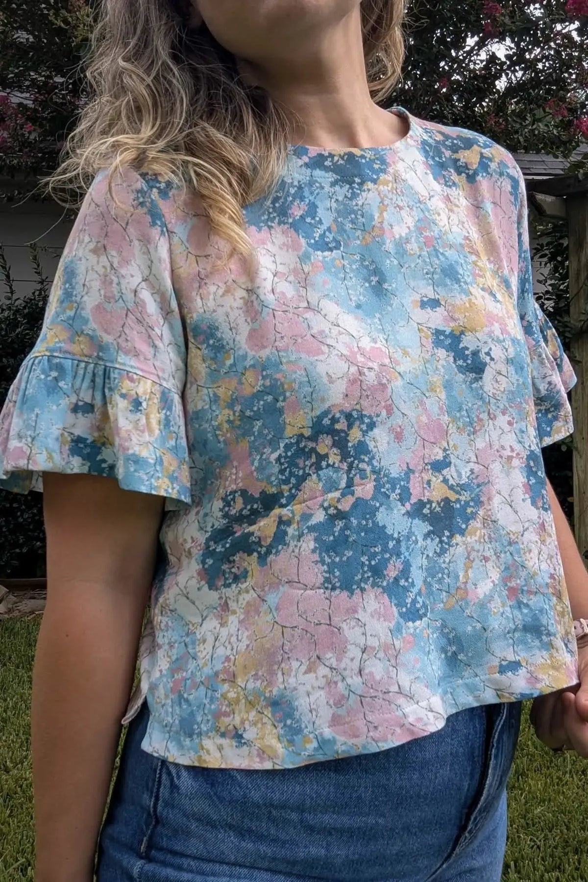 A woman wears a colorful, abstract-patterned top with ruffled sleeves and blue jeans.