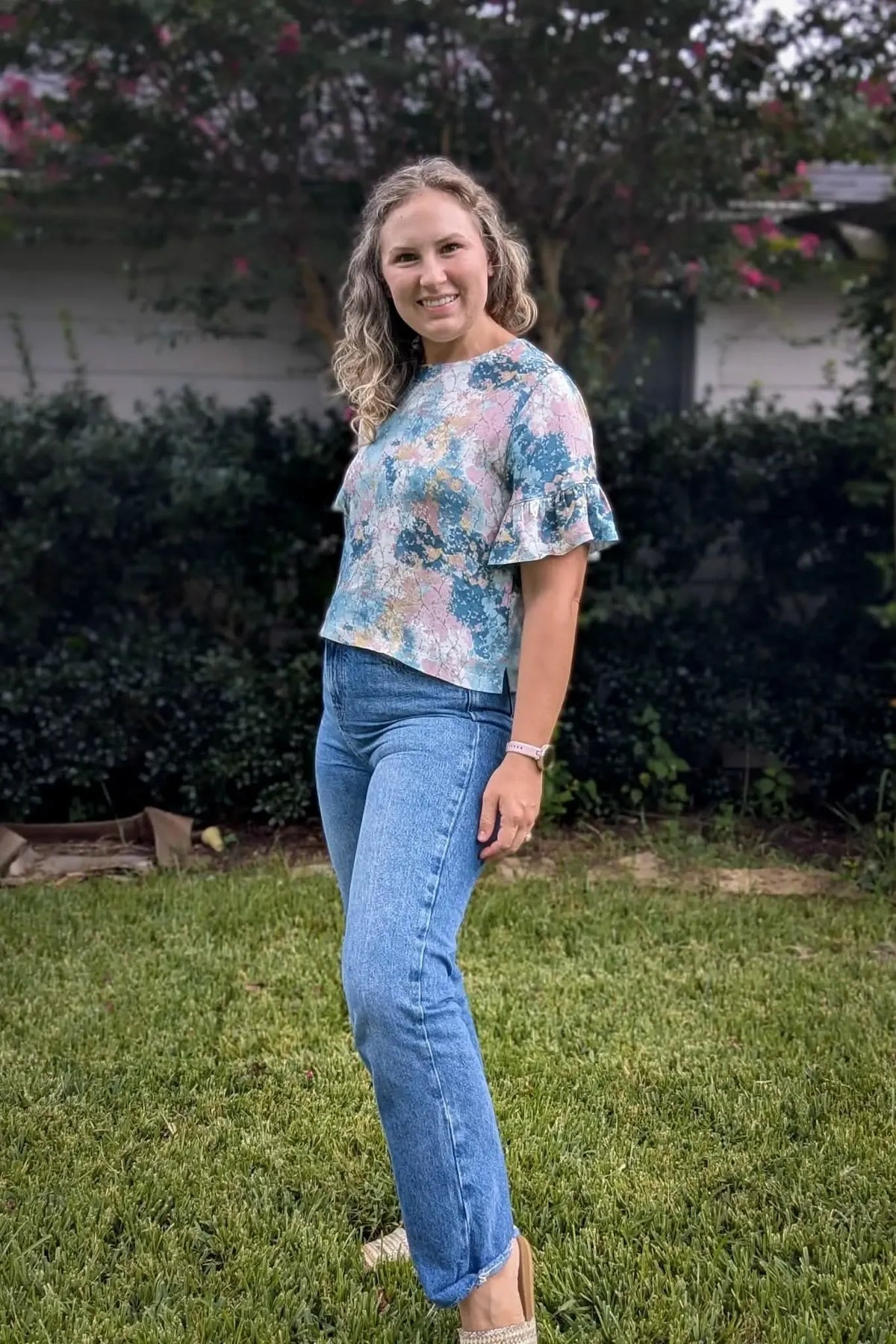 A woman in a floral top and jeans stands smiling in a grassy yard.