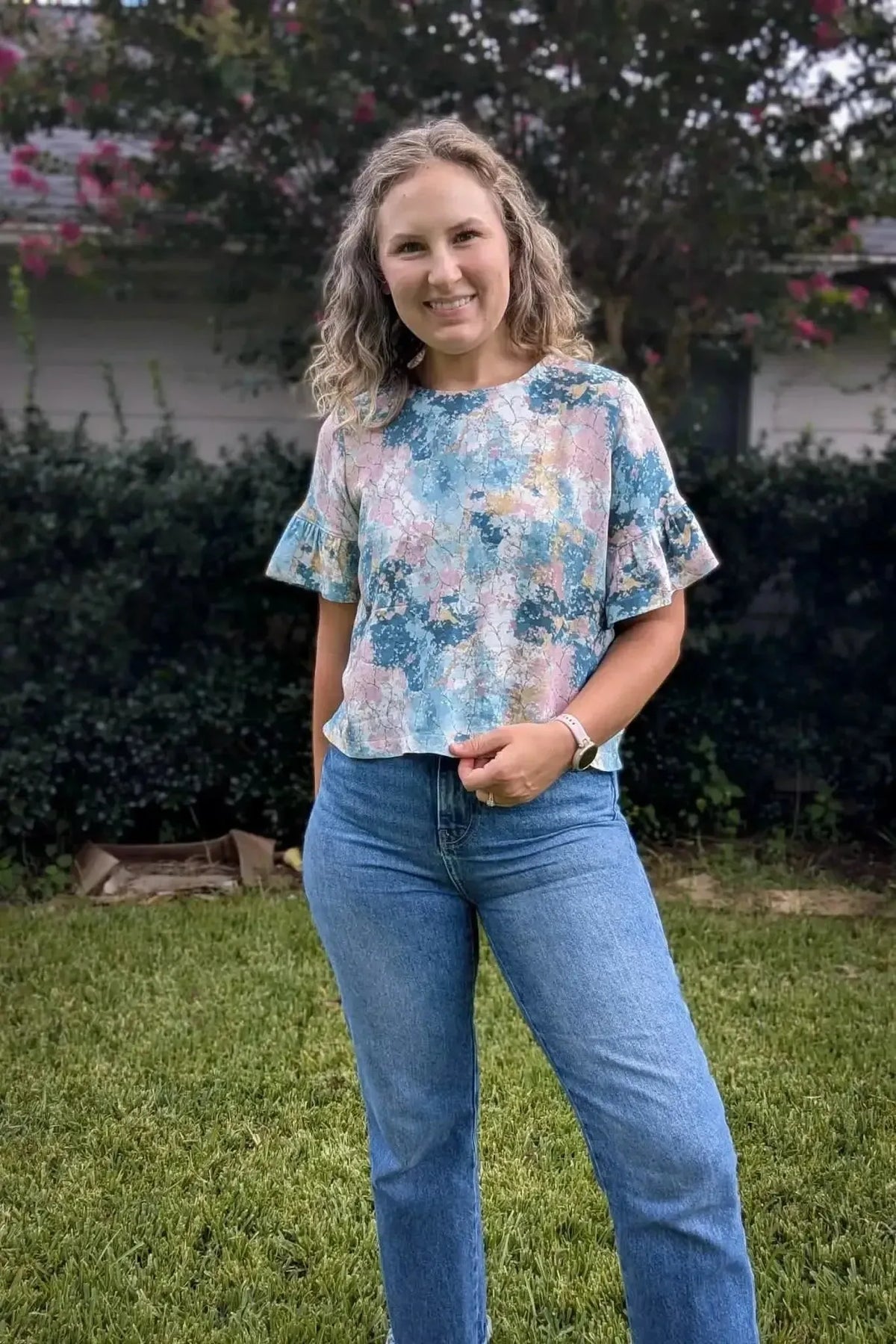 Woman modeling a short-sleeved blouse in abstract blue-pink floral fabric with jeans outdoors.